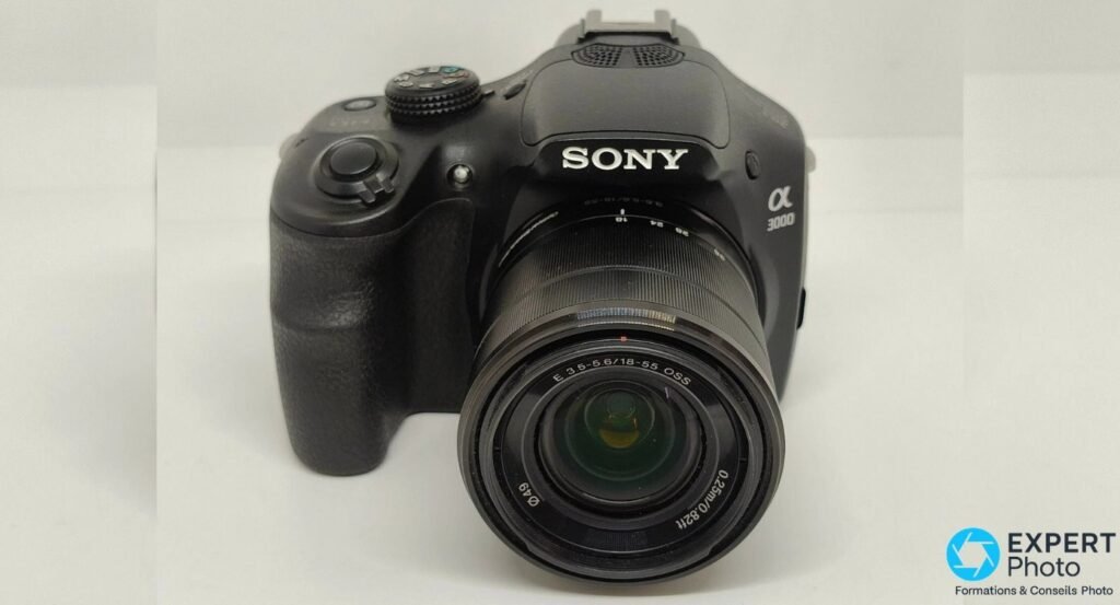 Vue frontale du Sony Alpha 3000 avec objectif Sony E 18-55 mm OSS rétracté, grip prononcé et logo Sony visibles. 