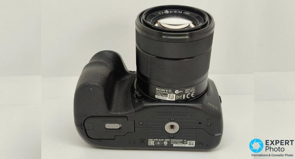 Vue de dessous du Sony Alpha 3000 avec étiquettes ILCE-3000 et SEL1855, filetage trépied 1/4 et trappe batterie.