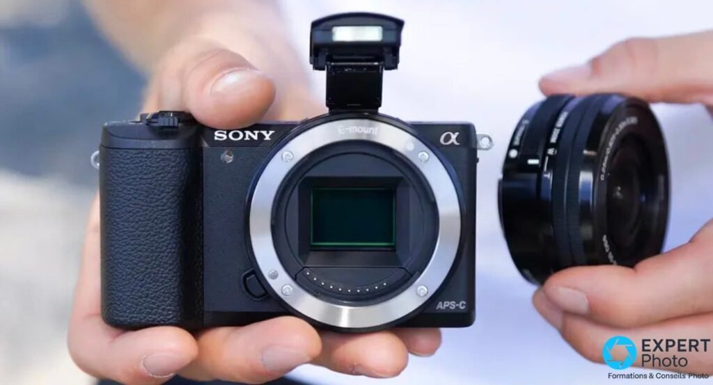 Sony Alpha 5100 tenu en main, capteur APS-C visible, flash intégré déployé, zoom Sony E 16-50 mm OSS détaché à droite.