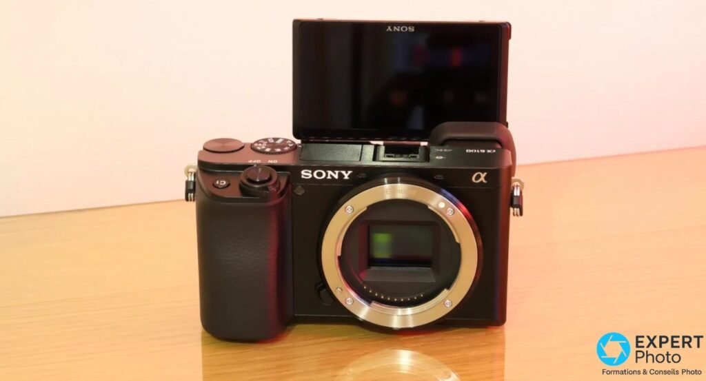 Sony Alpha 6100 boîtier nu vu de face, baïonnette E-mount et capteur APS-C visibles, écran rabattu vers le haut. 
