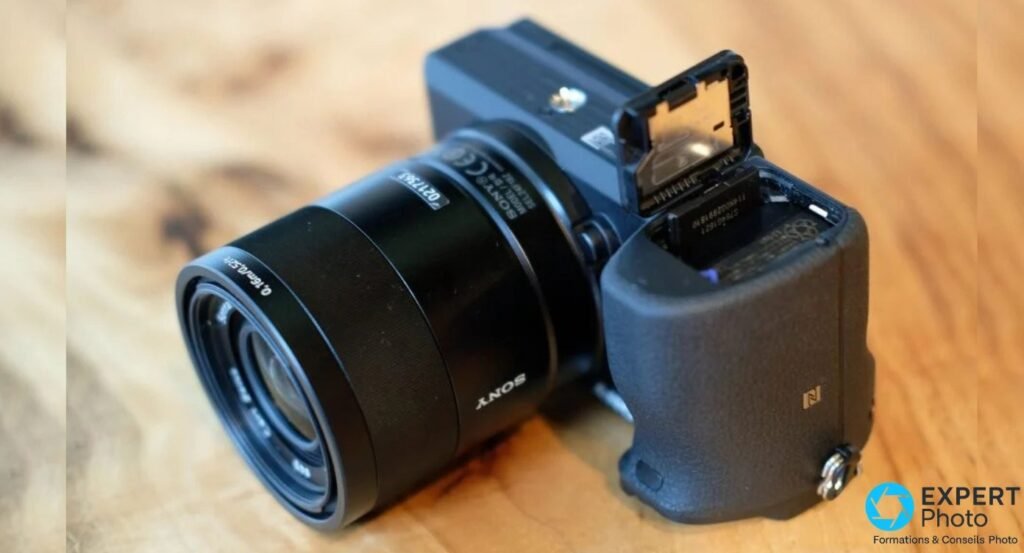 Sony Alpha 6500 vu de trois-quarts avant avec compartiment batterie NP-FW50 et carte SD ouvert sous le grip.