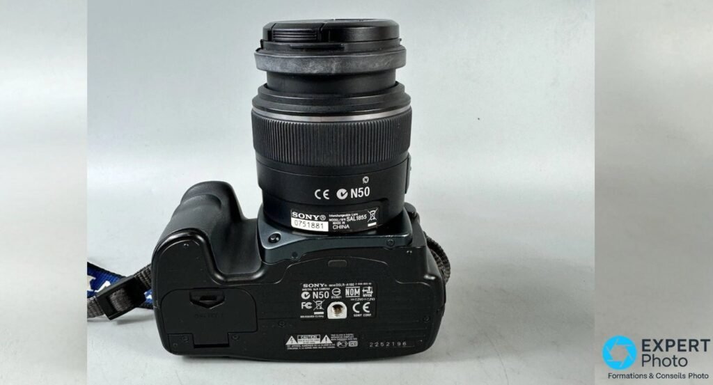 Vue de dessous du Sony Alpha 100 (DSLR-A100) avec étiquette modèle, numéro de série et marquages constructeur visibles.
