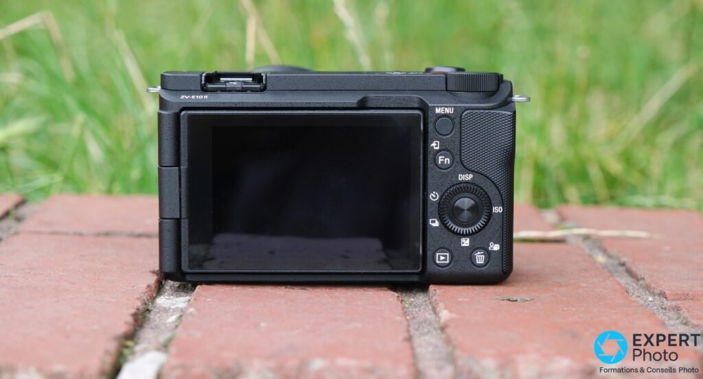  Dos du Sony ZV-E10 II noir : ecran, boutons MENU et Fn, molette de reglage, sur briques avec herbe en fond 