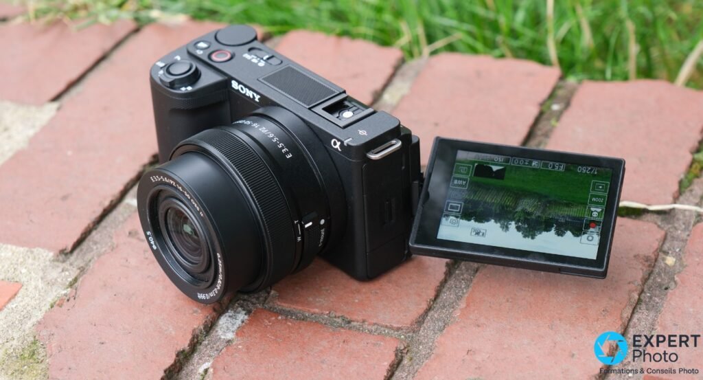 Sony ZV-E10 II noir de trois quarts avant, ecran orientable deploye, objectif PZ 16-50mm, sur briques rouges