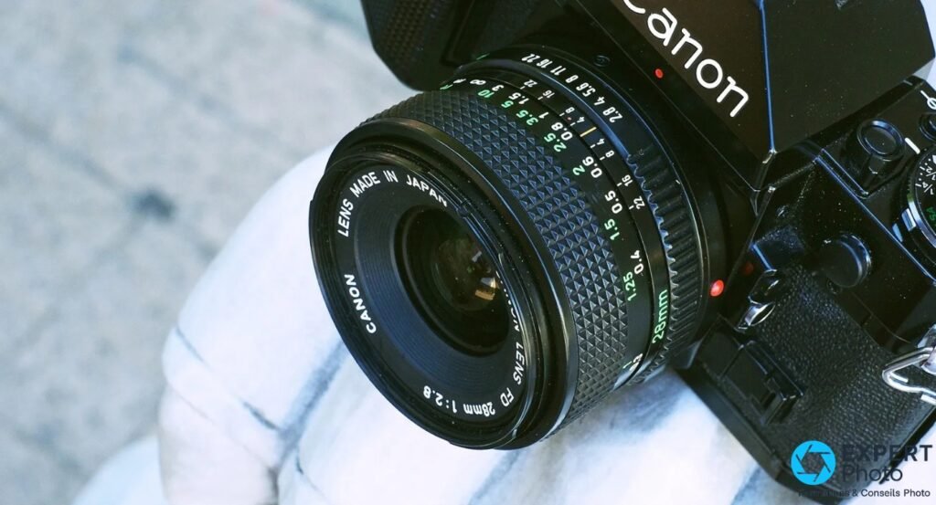 Canon FD 28mm f/2.8, face avant en plongée, inscription Canon FD 28mm 1:2.8 lisible sur l'anneau, boîtier Canon en arrière-plan