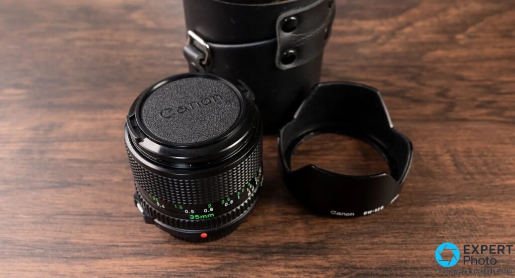  Canon New FD 35mm f/2 avec bouchon Canon, pare-soleil BW-52 et étui cuir d'origine posés sur bois.