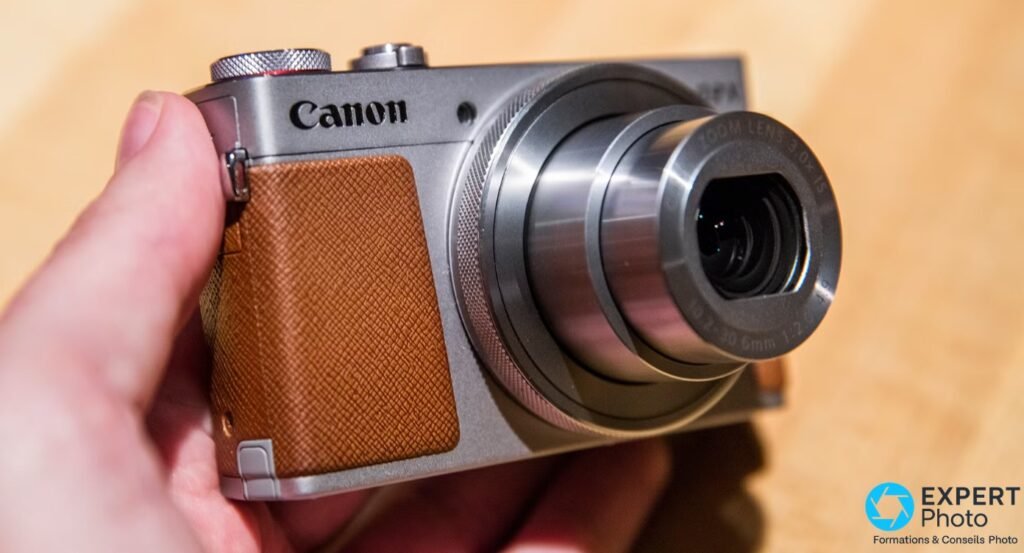 Canon PowerShot G9 X tenu en main, angle 3/4 avant gauche, zoom étendu, revêtement caramel et bague zoom moletée visibles