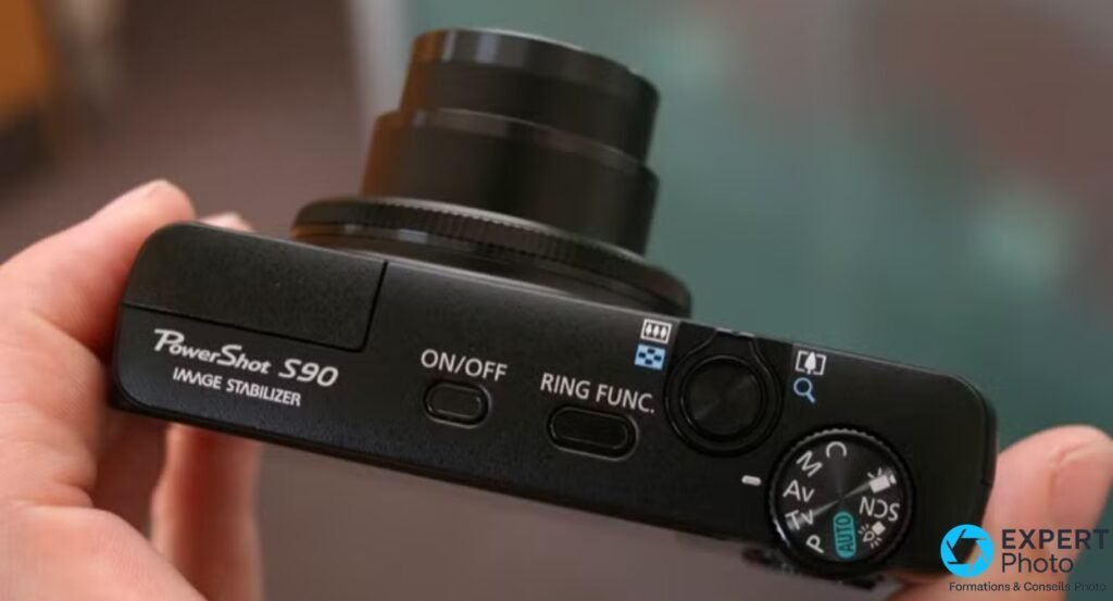 Canon PowerShot S90 vu du dessus tenu en main, boutons ON/OFF et RING FUNC., molette PASM et objectif sorti