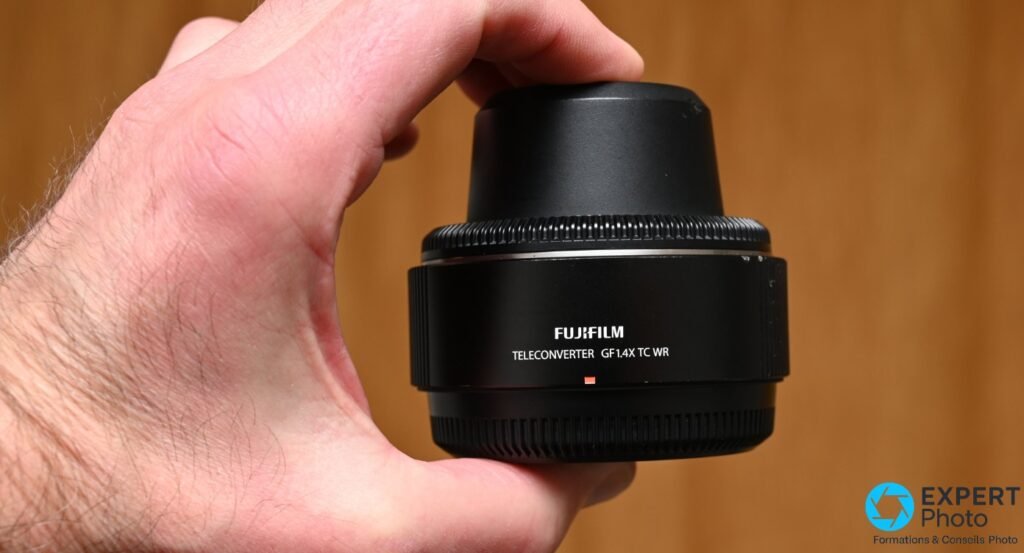 Téléconvertisseur Fujifilm GF 1.4X TC WR noir tenu en main, face avant avec capuchon, inscription TELECONVERTER GF1.4X TC WR lisible