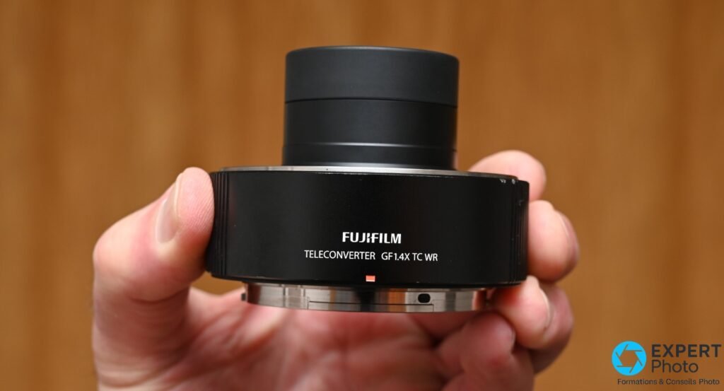 Fujifilm GF 1.4X TC WR présenté dans les deux mains, face avant avec inscription TELECONVERTER GF1.4X TC WR et repère rouge lisibles