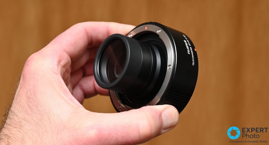Fujifilm GF 1.4X TC WR tenu en main, lentille frontale visible avec reflets bleutés du traitement antireflet et contacts baïonnette