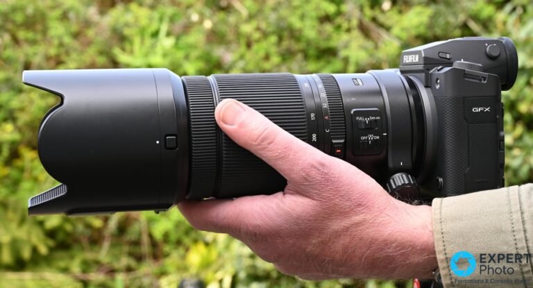 Objectif Fujinon GF 100-200mm f/5.6 avec pare-soleil monté sur boîtier Fujifilm GFX, tenu en main en extérieur, fond de feuillage flou