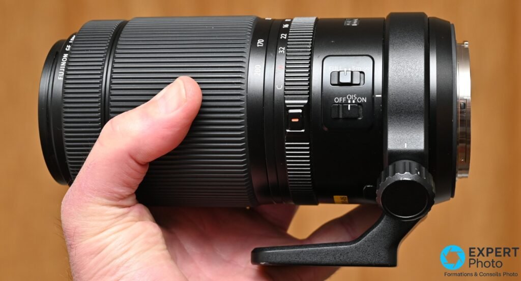  Objectif Fujinon GF 100-200mm f/5.6 tenu en main de profil, commutateur OIS OFF/ON et bagues de zoom et ouverture visibles