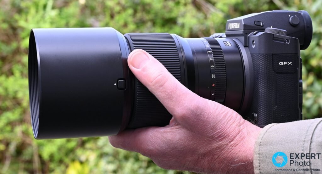 Fujinon GF 110mm f/2 avec pare-soleil monte sur boitier Fujifilm GFX, tenu en main, fond vegetal flou