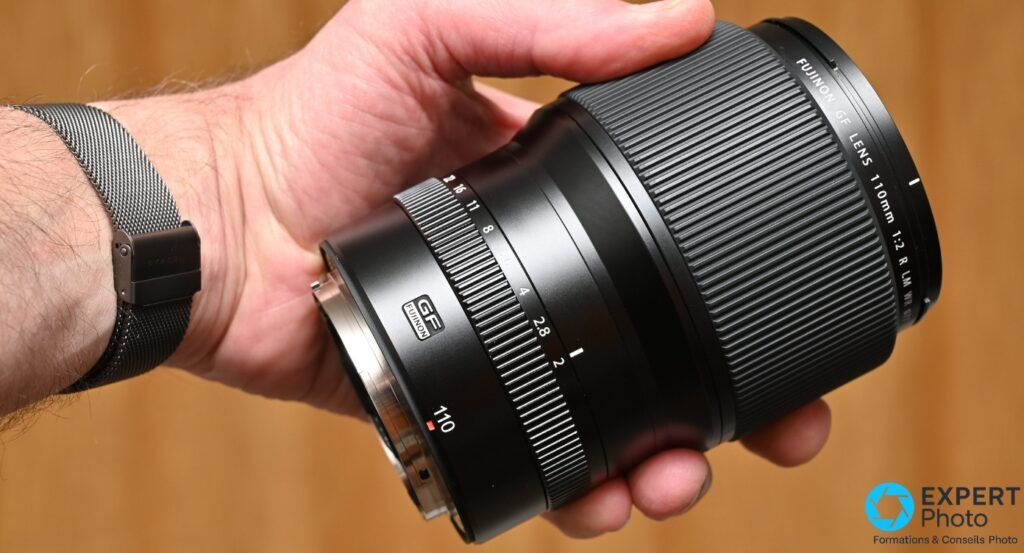 Objectif Fujinon GF 110mm f/2 en main, angle 3/4 avant, badge GF Fujinon et bague de diaphragme visibles