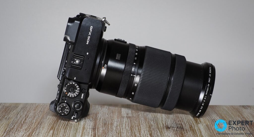 Fujifilm GF 45-100mm f/4 monté sur GFX 50R, profil gauche, objectif étendu, posé sur une surface en bois, fond gris.