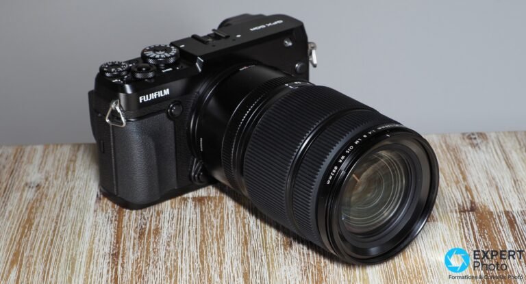 Avis Fujifilm GF 45-100mm f/4 : le zoom de travail des utilisateurs GFX Fujifilm GF 45-100mm f/4 sur GFX 50R, vue 3/4 avant, lentille frontale Ø82mm visible, posé sur bois, fond gris.