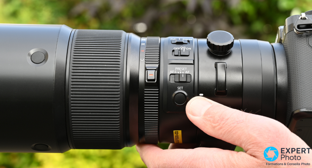 Commutateurs AF-L, PRESET AF et SET du Fujifilm GF 500mm f/5.6 vue profil, main en prise sur l'objectif 