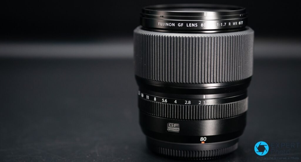 Fujifilm GF 80mm f/1.7 R WR posé sur fond anthracite, bague avant avec inscription « FUJINON GF LENS 80mm 1:1.7 R WR Ø77 » lisible