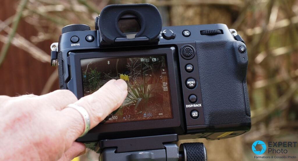  Fujifilm GFX 100S dos, écran tactile en live view avec scène florale, main pointant vers l'écran, boutons AEL et MENU/OK visibles