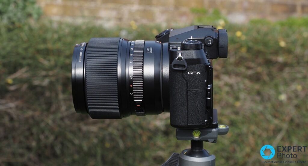 Fujifilm GFX 100S avec objectif Fujinon GF vu de profil droit, sur trépied, fond herbe et mur flous