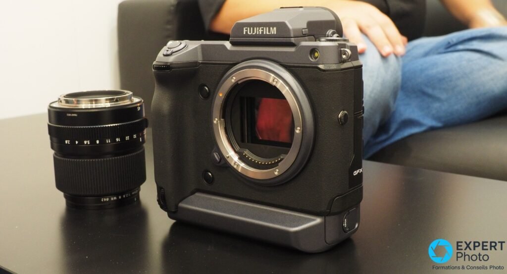 Fujifilm GFX 100 boîtier nu, monture G ouverte avec reflet rougeâtre, objectif GF posé à gauche sur surface noire 