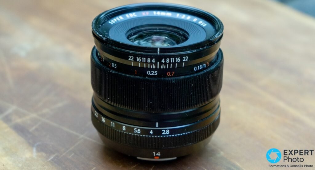 Fujifilm XF 14mm f/2.8 R noir posé sur bois, bague diaphragme f/2.8–22 et bague MAP visibles, sans capuchon