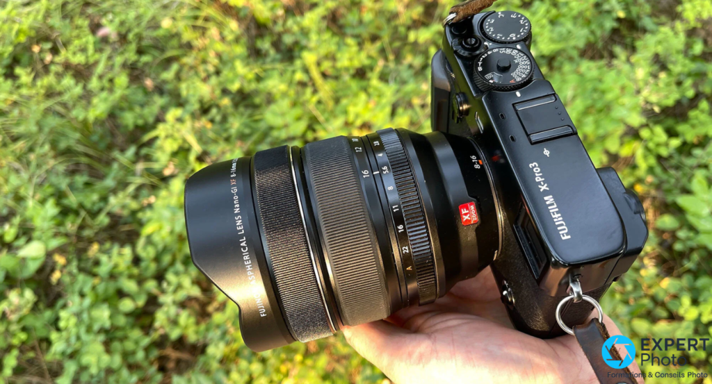  Fujifilm XF 8-16mm f/2.8 R LM WR monté sur boîtier Fujifilm X-Pro3, tenu en main par-dessus, fond végétal vert