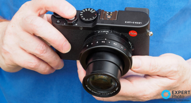Leica D-Lux 8 : avis complet sur le compact expert zoom pour voyager léger Leica D-Lux 8 noir tenu à deux mains, objectif DC Vario-Summilux f/1,7 rétracté, bague d'ouverture visible, fond bleu flou