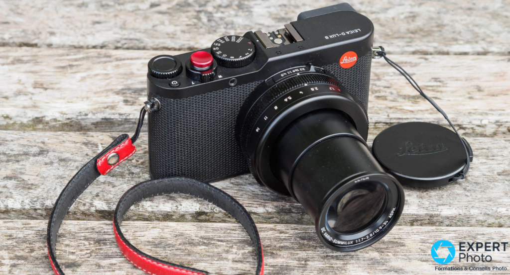Leica D-Lux 8 noir, zoom DC Vario-Summilux étendu, cache objectif Leica et sangle cuir rouge posés, surface bois gris