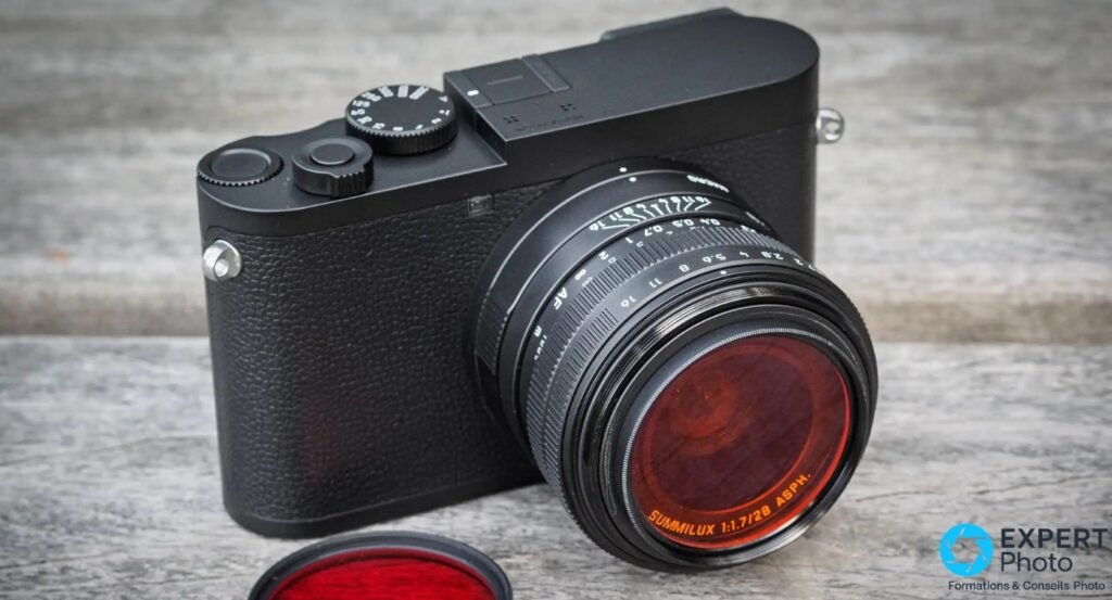 Leica Q2 Monochrom angle 3/4 avant avec filtre rouge monté sur le Summilux 28 mm, capuchon rouge au premier plan.