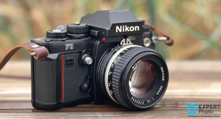 Nikkor 50mm f/1.4 AI-S : pour les photographes Nikon F à l’aise avec le manuel Nikkor 50mm f/1.4 monté sur reflex argentique Nikon F3 noir, vue 3/4 avant, surface bois, fond bokeh vert
