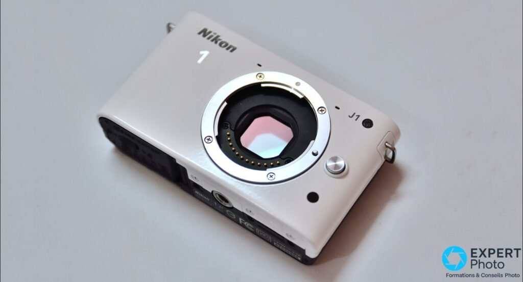 Nikon 1 J1 blanc seul en plongée, monture ronde exposée avec contacts dorés visibles, fond gris clair