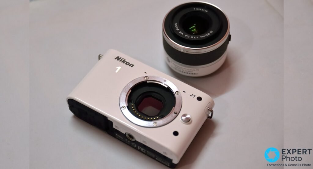 Nikon 1 J1 blanc posé en biais, monture exposée sans objectif, zoom 1 NIKKOR 10-30mm déposé à côté, fond gris