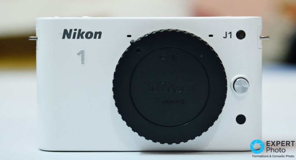 Nikon 1 J1 blanc sans objectif, cache-objectif noir marqué Nikon 1 mount monté, vue frontale sur fond blanc