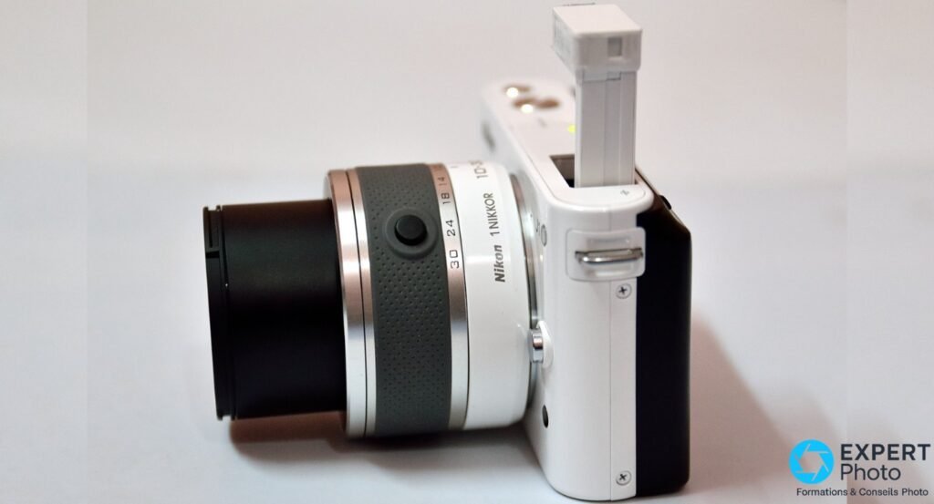 Nikon 1 J1 blanc de profil gauche, zoom 1 NIKKOR 10-30mm étendu, flash intégré sorti et relevé, fond blanc