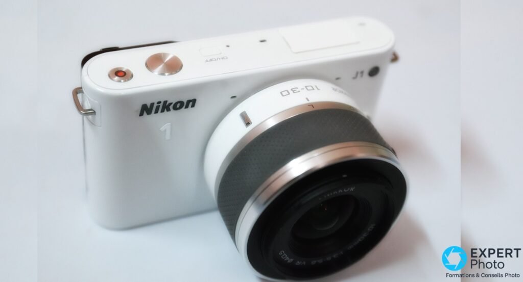 Nikon 1 J1 blanc avec zoom 1 NIKKOR 10-30mm monté et déployé, angle 3/4 avant, fond blanc homogène