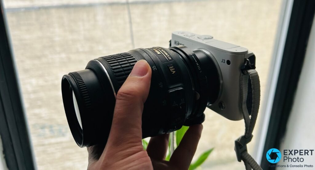 Nikon 1 J3 argenté vu de profil, tenu en main avec un zoom Nikkor AF-S monté via adaptateur, devant fenêtre