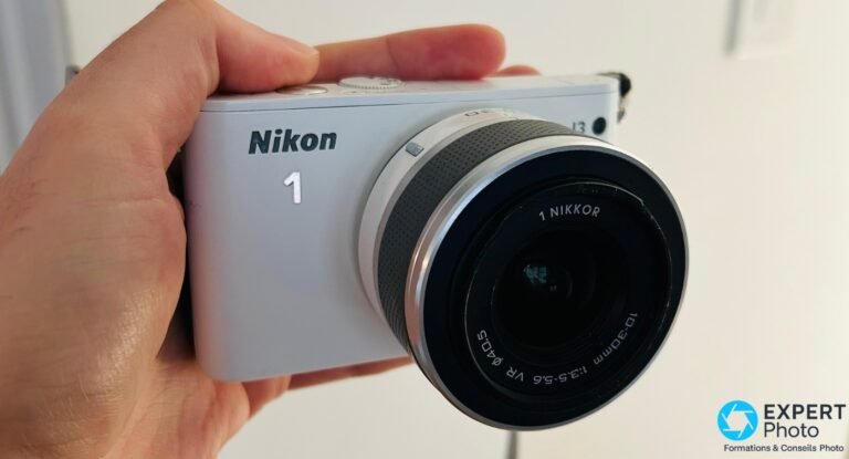 Nikon 1 J3 : ce qu’il est vraiment en 2026 sur le marché de l’occasion Nikon 1 J3 blanc tenu en main, vue face avant avec zoom 1 Nikkor 10-30mm f/3.5-5.6 VR monté, fond clair