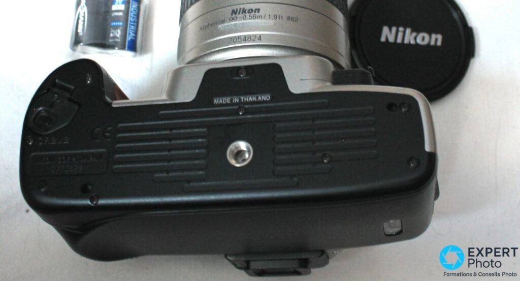 Dessous du Nikon F65, mention MADE IN THAILAND, vis de pied, objectif serie 2054824, pile et bouchon Nikon a cote.