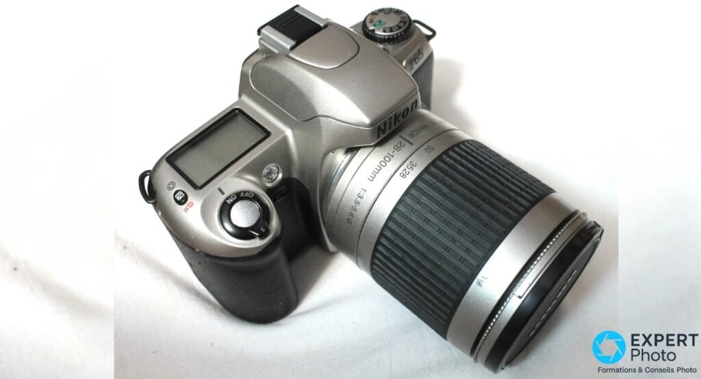 Nikon F65 argente, vue dessus-dos 3/4, objectif 28-100 mm en position courte, afficheur LCD dos visible, flash replie.