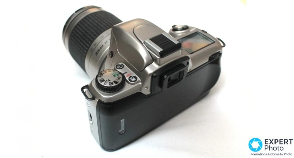 Nikon F65 argente, vue de dessus, molette de modes P A S M et scenes lisible, viseur oculaire et objectif zoom visibles.