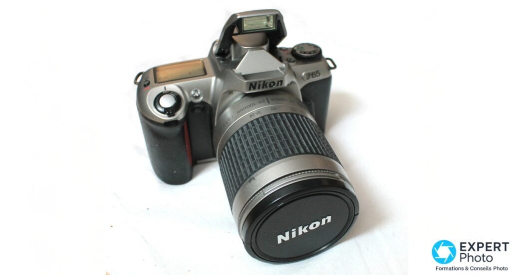 Nikon F65 argente, vue 3/4 avant, flash integre releve, zoom Nikon 28-100 mm monte avec bouchon avant, fond blanc.
