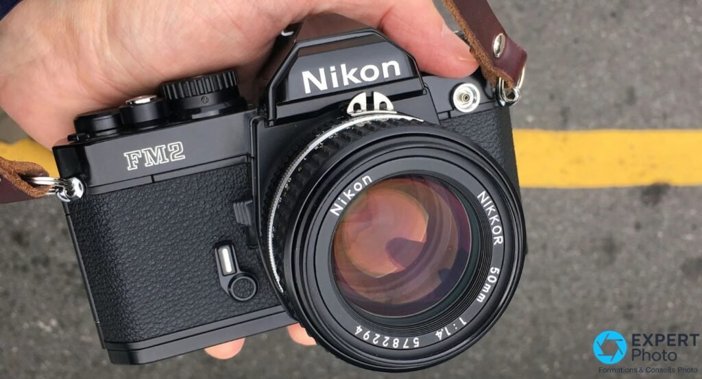 Nikon FM2n noir tenu en main, face 3/4 avant, objectif Nikon Nikkor 50mm f/1.4, sangle cuir, fond asphalte