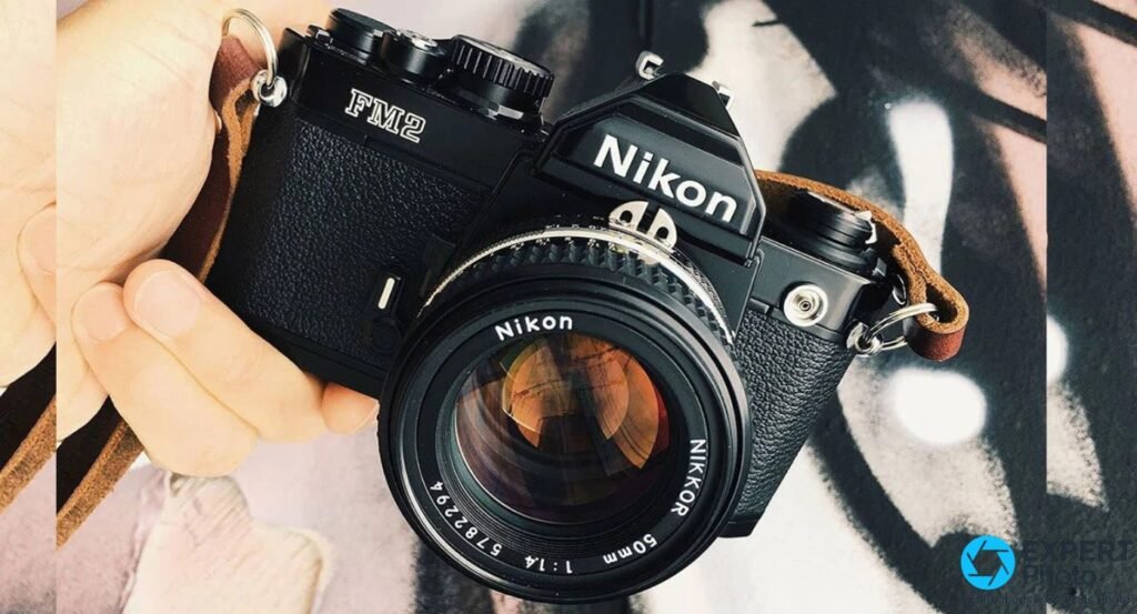 Nikon FM2n noir vu en plongée, tenu en main, objectif Nikon Nikkor 50mm f/1.4, sangle cuir brun, fond abstrait