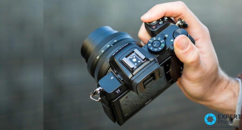 Nikon Z50 tenu en main, vue dessus, molette modes A/S/P/M/AUTO visible, griffe flash ouverte, objectif 16-50mm monté, fond gris flou