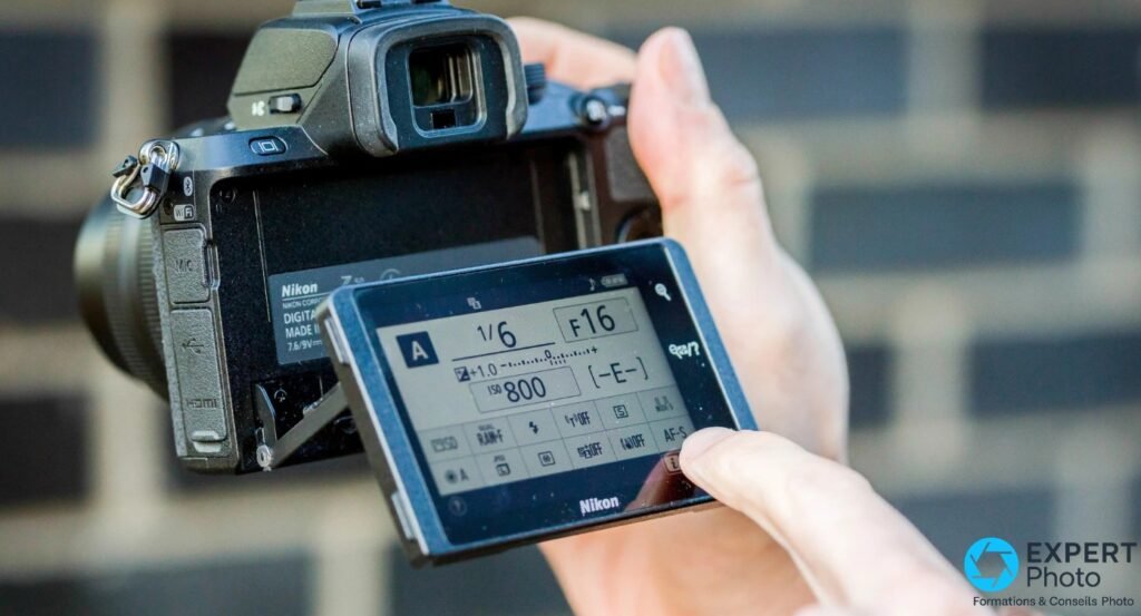 Nikon Z50 tenu à deux mains, écran déplié vers le bas affichant menu réglage rapide mode A, 1/6, F16, ISO 800, fond extérieur brique
