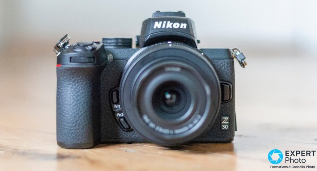Nikon Z50 noir de face, objectif zoom monté, logo Z50 visible bas droite, posé sur surface bois, fond gris clair flou