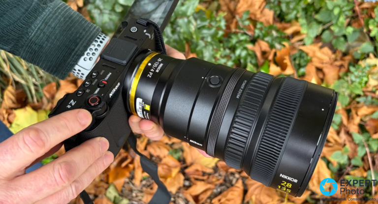 Nikon ZR noir tenu à deux mains, objectif NIKKOR Z 28-135/4 PZ monté, bague dorée visible, fond de feuilles mortes