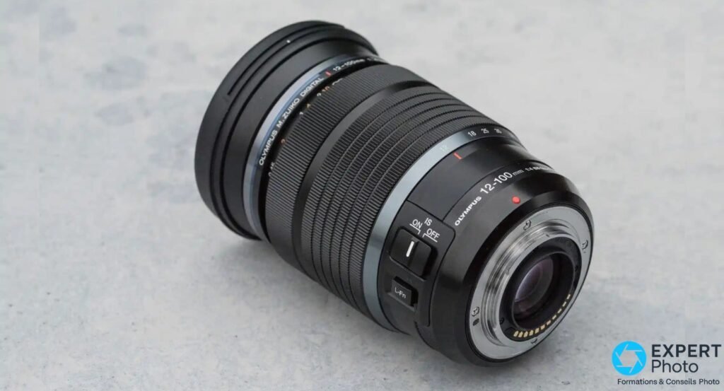 M.Zuiko 12-100mm f/4 IS PRO vu de 3/4 arrière, commutateur IS ON/OFF et bouton L-Fn visibles, baïonnette Micro 4/3 exposée